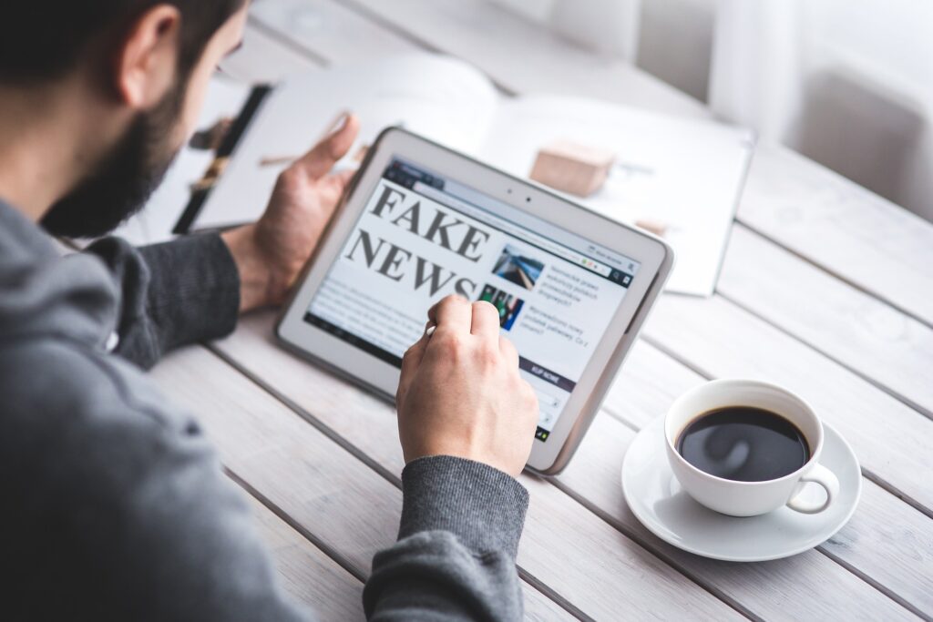 Mann tippt auf einem Tablet mit einem "Fake News"-Artikel vor erhelltem Holztisch mit Kaffetasse und offenem Heft