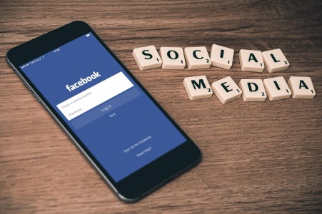 Ein Handy, auf dem Facebook geöffnet ist, liegt auf einem Tisch. Daneben ist mit Spielsteinen der Schriftzug "Social Media" gelegt.
